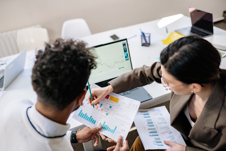 Two colleagues in a meeting room discussing financial charts and graphs on a laptop and paper.